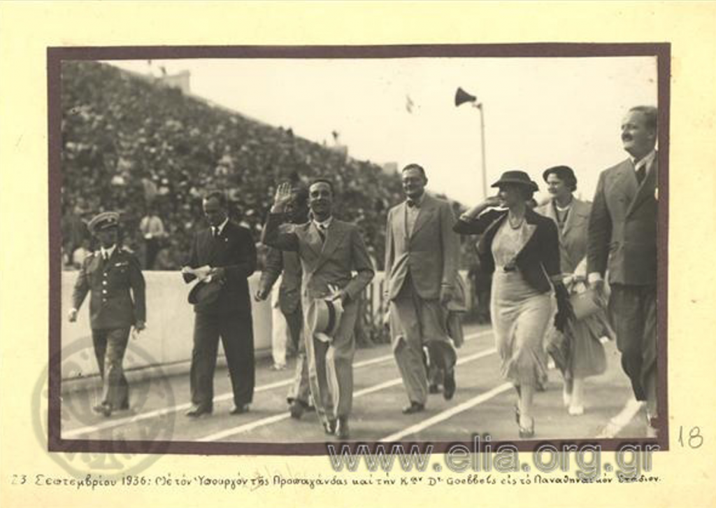 Ο δήμαρχος Αθηναίων Κώστας Κοτζιάς (άκρη δεξιά) με τον υπουργό προπαγάνδας της ναζιστικής Γερμανίας Josef Goebbels, ο οποίος χαιρετά το πλήθος στο Παναθηναϊκό Στάδιο με αφορμή τους Ολυμπιακούς Αγώνες του 1936. 